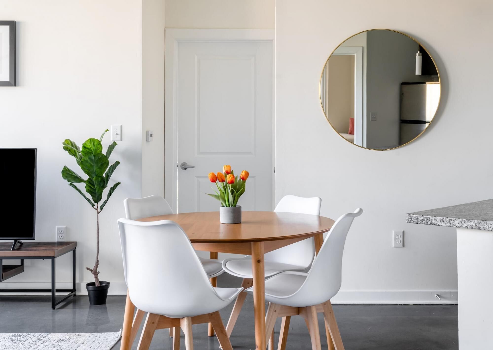 A dining area with a round wooden table, white chairs, a potted plant, and a flat-screen TV.