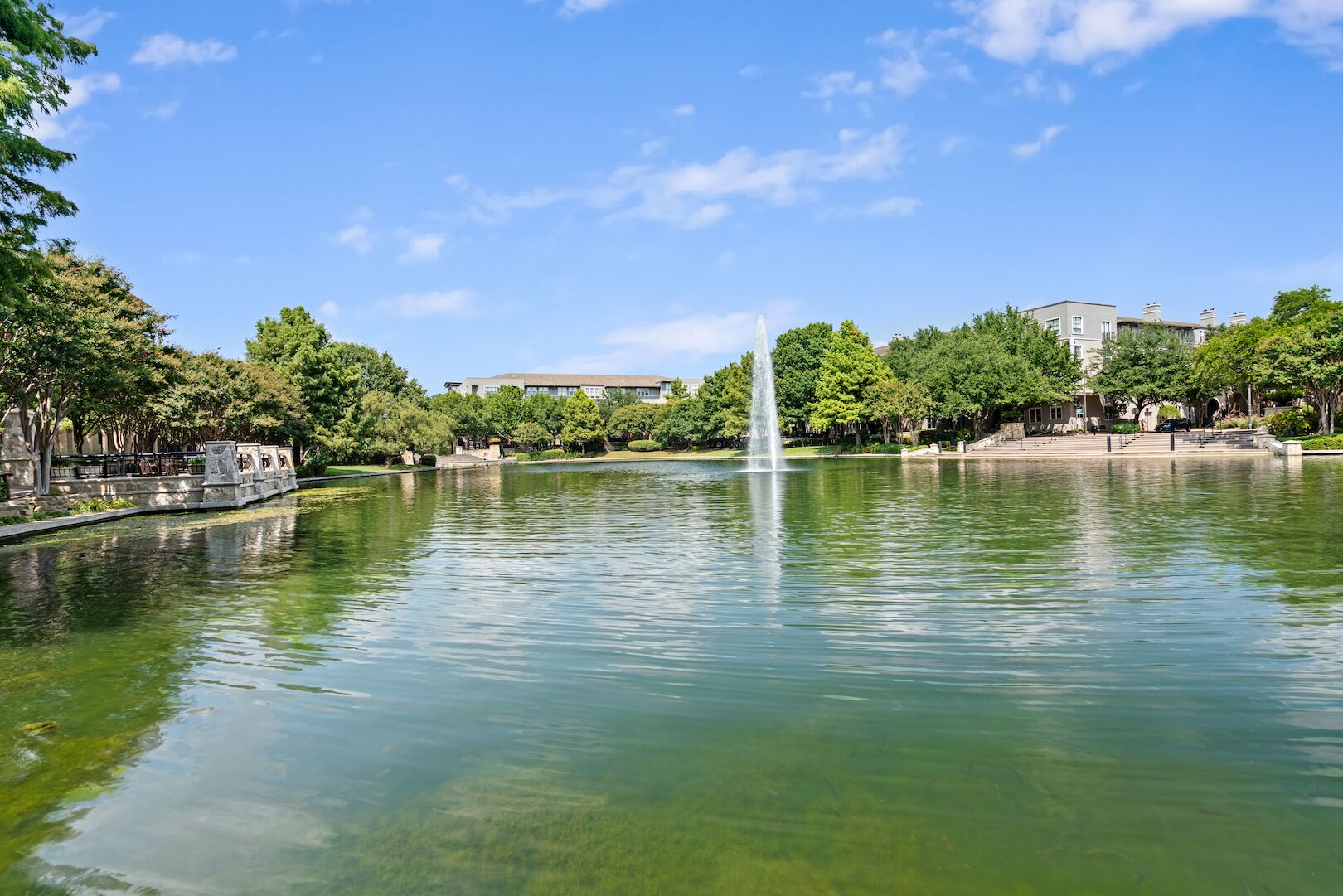 Un lago tranquilo con una fuente central, rodeado de árboles y edificios.