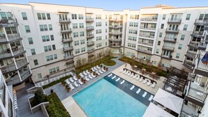 A modern apartment building with a central courtyard featuring a swimming pool, lounge chairs, and potted plants.