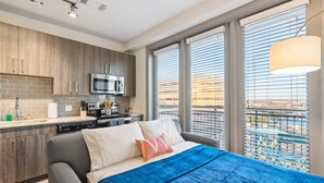 A modern bedroom with a bed, a view of the city through large windows, and a kitchenette with a microwave and stove.