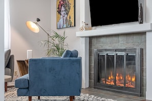 Een woonkamer met een blauwe bank, een open haard, een televisie en een schilderij aan de muur.