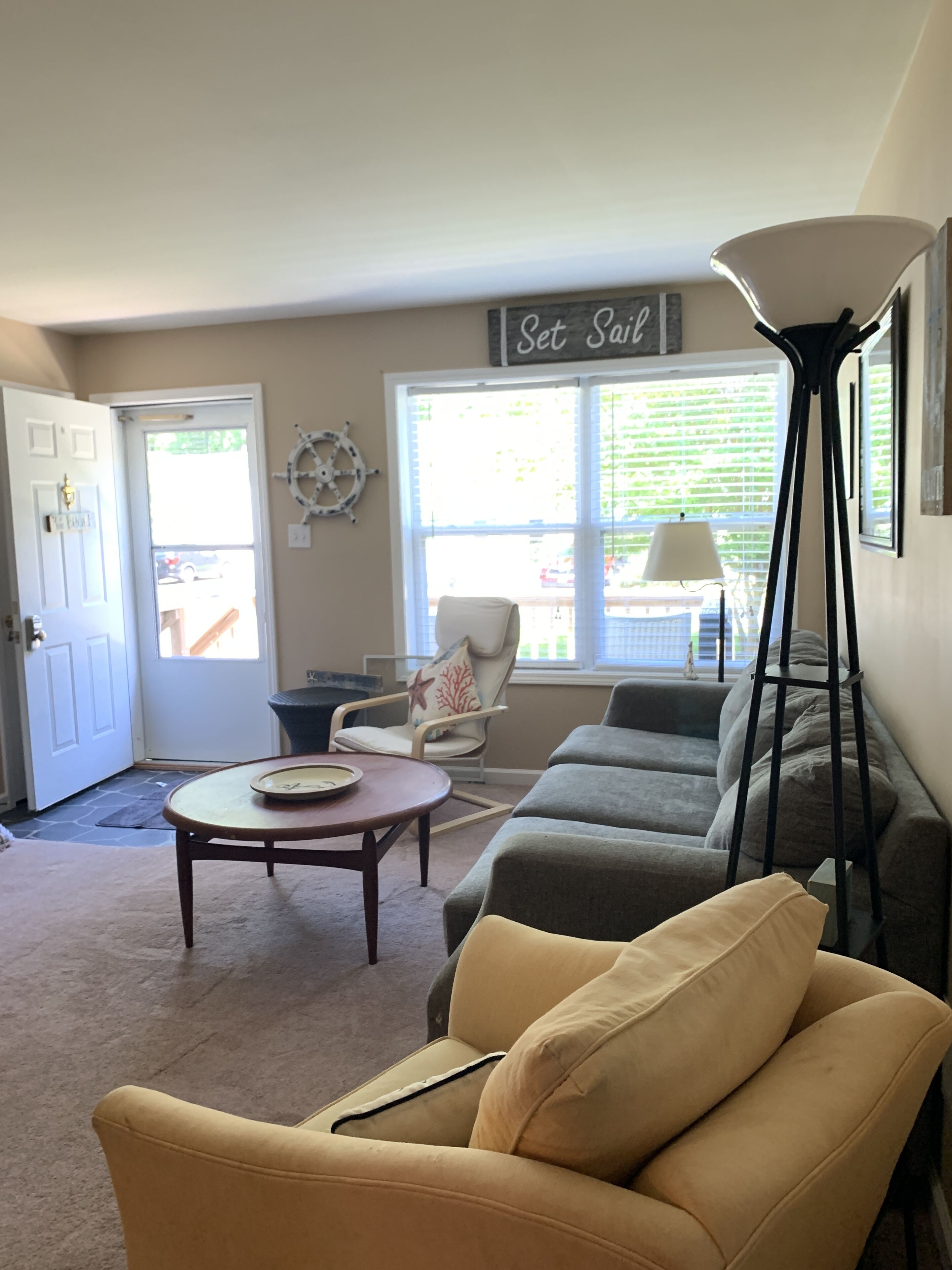 A living room with a yellow armchair, a gray sofa, a white chair with a star pillow, a round coffee table, and a wall-mounted sign that reads "Set Sail".