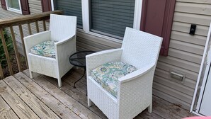 Zwei weiße Korbstühle mit gemusterten Kissen auf einer Holzterrasse, ein kleiner runder Tisch und ein Haus mit beige coloren Anstrich und braunen Fensterläden.