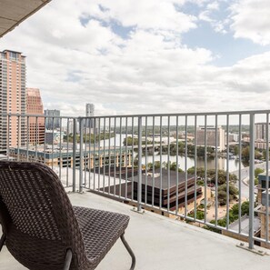 A balcony with a wicker chair, overlooking a cityscape with buildings and a river.