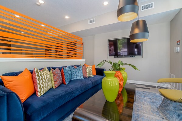 A modern living room with a blue sofa, colorful cushions, a green vase, and a television on the wall.