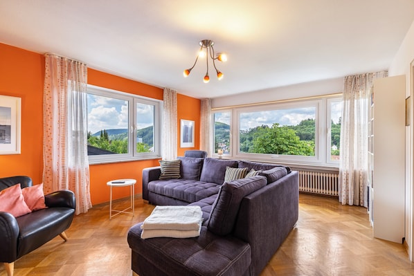 A living room with a sofa, a chair, a small table, and large windows with a view of greenery.