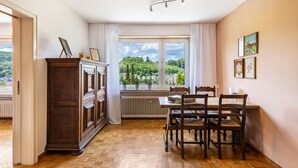 Habitació amb sòl de fusta, taula de menjador amb cadires, radiador i finestra amb vista a vegetació.