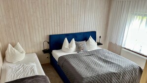 Ein Schlafzimmer mit blauem Kopfteil, zwei Betten, einem Nachttisch mit Lampe und einem Fenster mit Vorhängen.