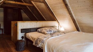 Ein Schlafzimmer mit schrägen Wänden, Holzbalken, einem Bett mit gemusterten Textilien und einem kleinen Hocker.