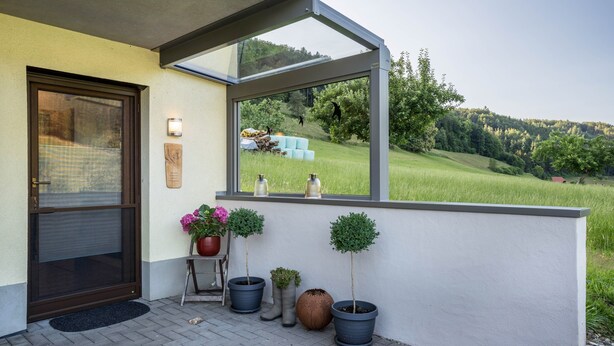 Een huisentree met een glazen deur, plantenpotten en uitzicht op een groen landschap.