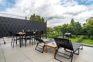 Een terras buiten met een tafel, stoelen, een barbecue en uitzicht op een tuin.