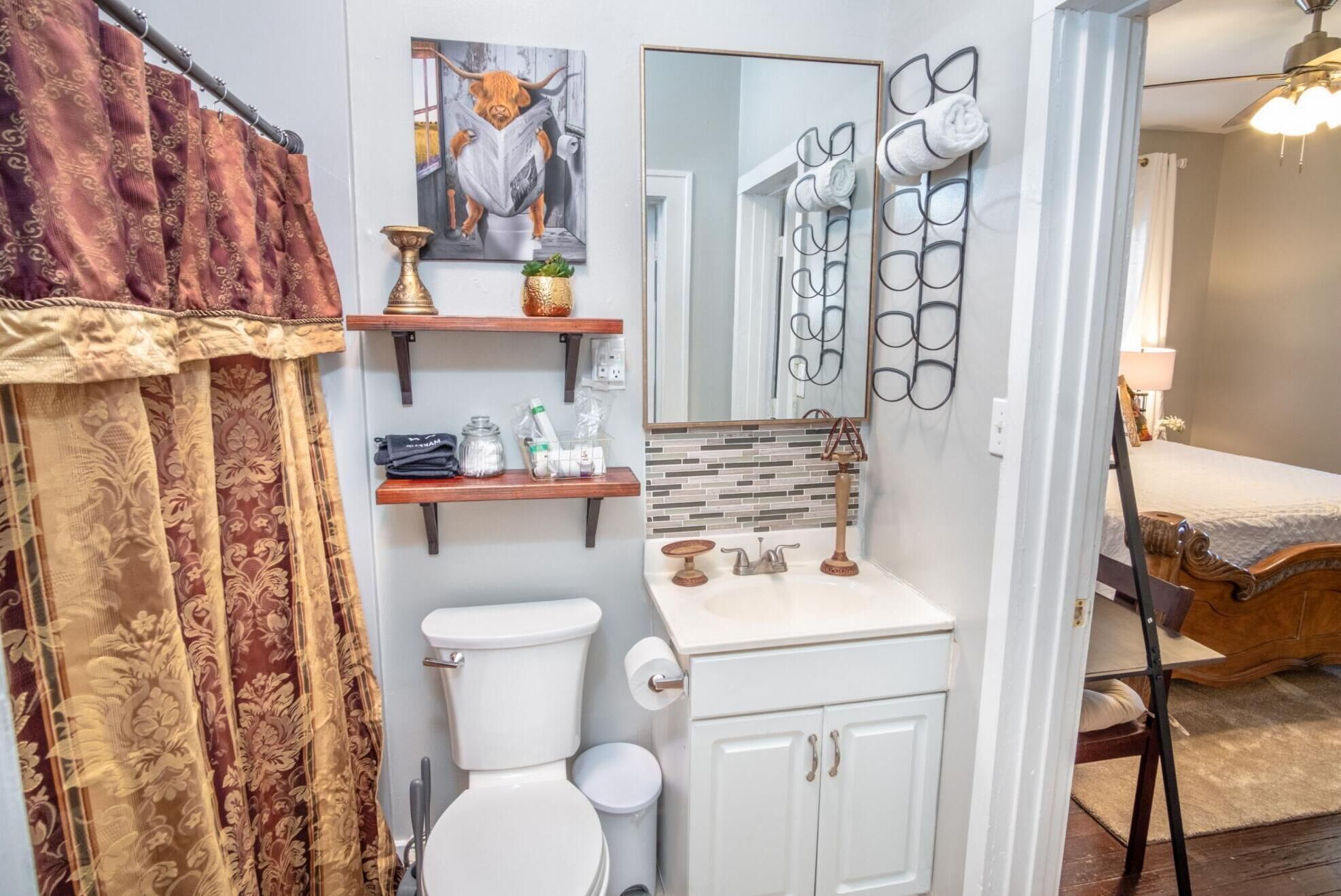 A bathroom with a toilet, sink, and mirror. There are shelves with toiletries and a painting of a bull.