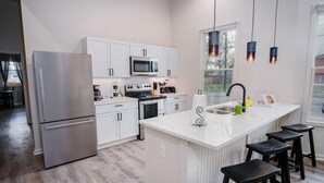 A modern kitchen with white cabinetry, a stainless steel refrigerator, a microwave, and a large island with bar stools. - Magnificent 4BR Near Famous Riverwalk and Downtown (San Antonio)