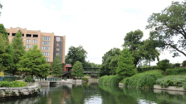 A serene riverside setting with a bridge, lush greenery, and a building with 'EM' signage. - Exceptional 5BR So Close to Riverwalk and Downtown (San Antonio)