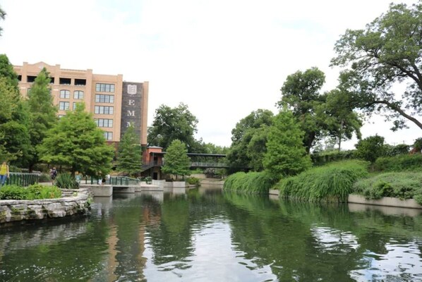 A serene riverside setting with a bridge, lush greenery, and a building with 'EM' signage. - Exceptional 5BR So Close to Riverwalk and Downtown (San Antonio)