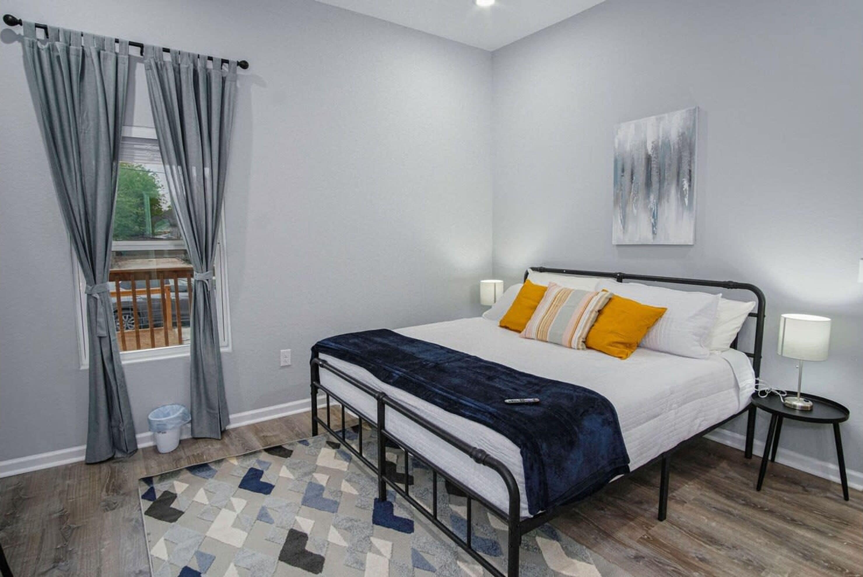 A bedroom with a bed, a window with curtains, a painting on the wall, and a small bedside table with a lamp.