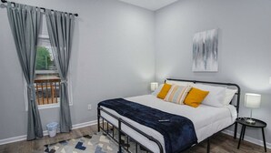 A bedroom with a bed, a window with curtains, a painting on the wall, and a small bedside table with a lamp. - Exceptional 5BR So Close to Riverwalk and Downtown (San Antonio)