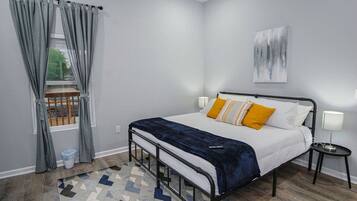 A bedroom with a bed, a window with curtains, a painting on the wall, and a small bedside table with a lamp.
