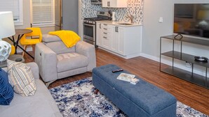 A compact living space with a kitchenette, a grey sofa, a blue ottoman, and a TV on a stand. - Remodeled Modern 1BR 1BA Apt Near Downtown 5min Pearl (San Antonio)