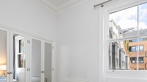 A hotel room with a large bed, a window with a view of buildings, a radiator, and a wardrobe. - The Belgravia Suite Next to Sloane Square (London)