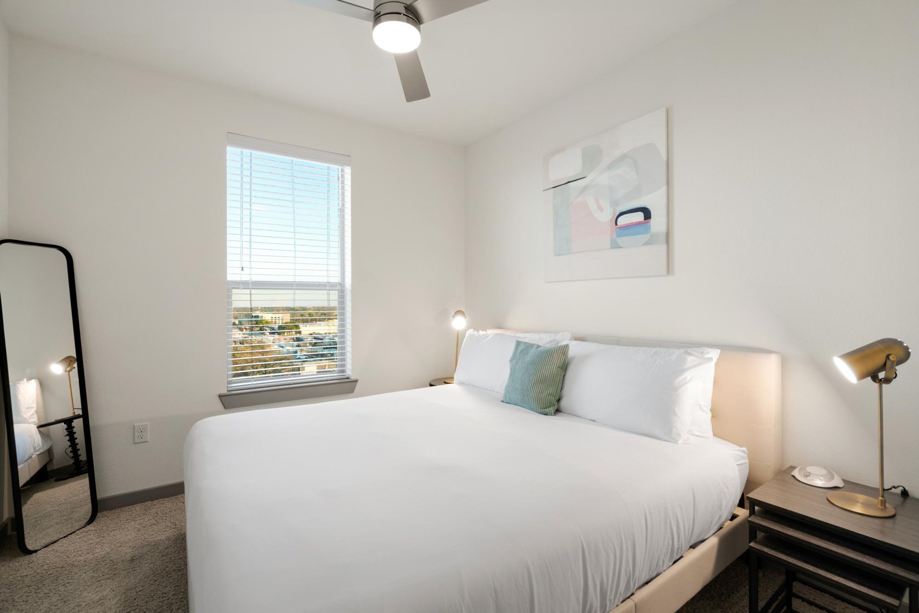 A bedroom with a large bed, a ceiling fan, a window with blinds, a bedside table with a lamp, and a framed picture on the wall.