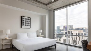 A modern hotel room with a large bed, two bedside tables with lamps, a framed picture, and a city view through large windows. - Austin Remarkable 1BD 1BA 2nd Street District Apartment (Austin)
