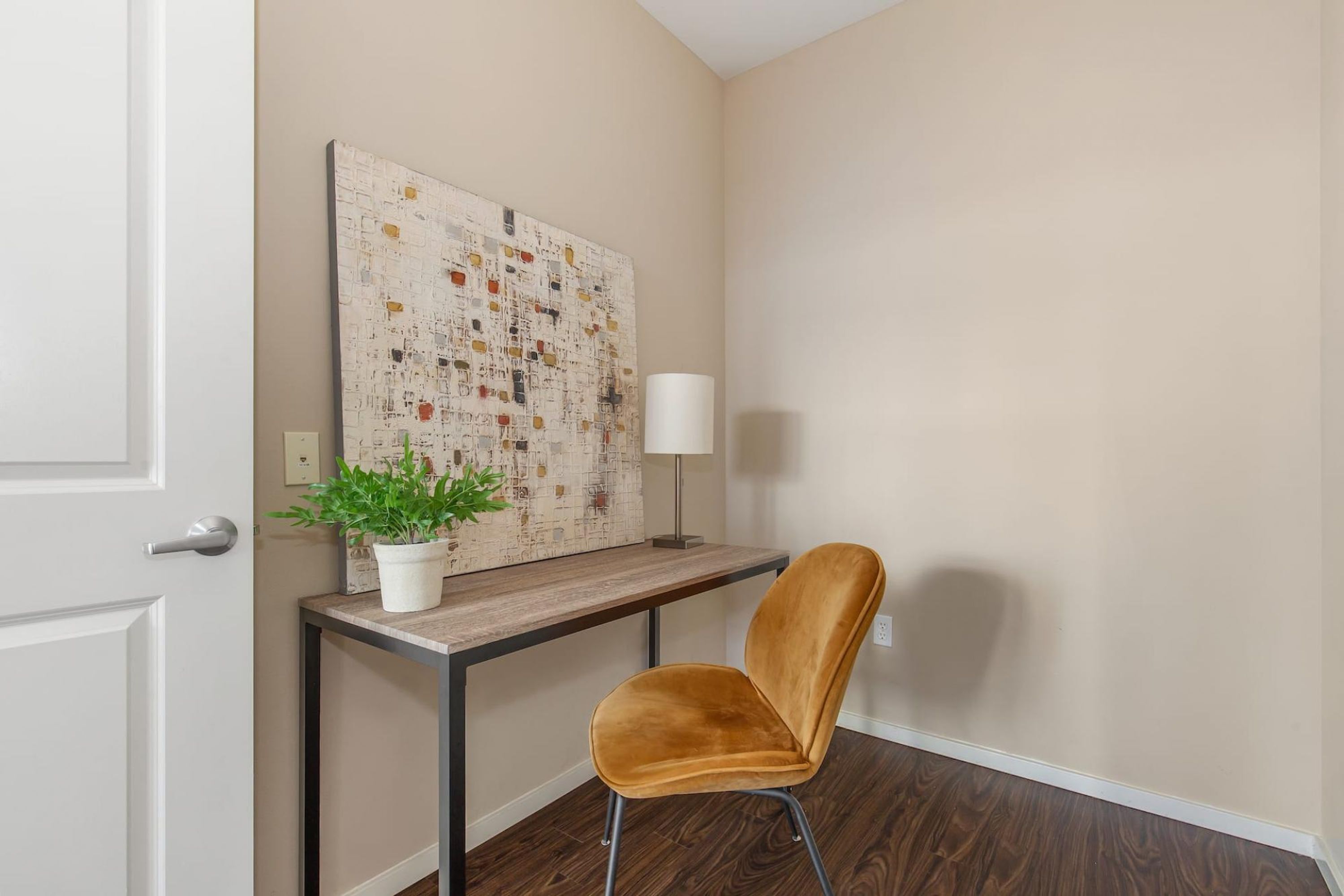 A room with a wooden desk, a chair, a potted plant, a lamp, and a large abstract painting on the wall.