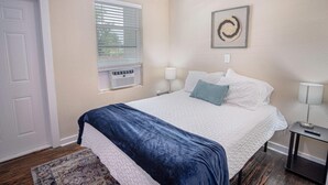 A bedroom with a bed, a window with blinds, a wall-mounted artwork, and a bedside table with a lamp.