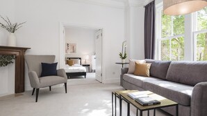 A modern living room with a grey sofa, a striped armchair, a small coffee table with a book, and a large window with curtains. - The King s Road Suite Next to Sloane Square (London)