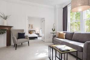 A modern living room with a grey sofa, a striped armchair, a small coffee table with a book, and a large window with curtains. - The King s Road Suite Next to Sloane Square (London)