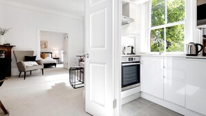 A modern kitchen with white cabinetry, a built-in oven, and a window with a view of greenery. - The King s Road Suite Next to Sloane Square (London)
