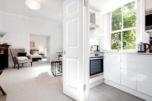 A modern kitchen with white cabinetry, a built-in oven, and a window with a view of greenery. - The King s Road Suite Next to Sloane Square (London)