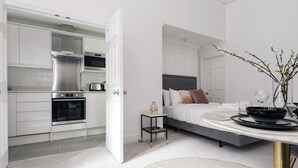 Une chambre d’hôtel moderne dotée d’une kitchenette, d’un lit avec une tête de lit et d’un coin repas avec une table dressée pour deux personnes.