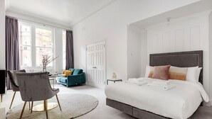 Une chambre moderne dotée d’un grand lit, d’un coin salon avec un fauteuil et une petite table, et offrant une vue sur la ville grâce à de grandes fenêtres.