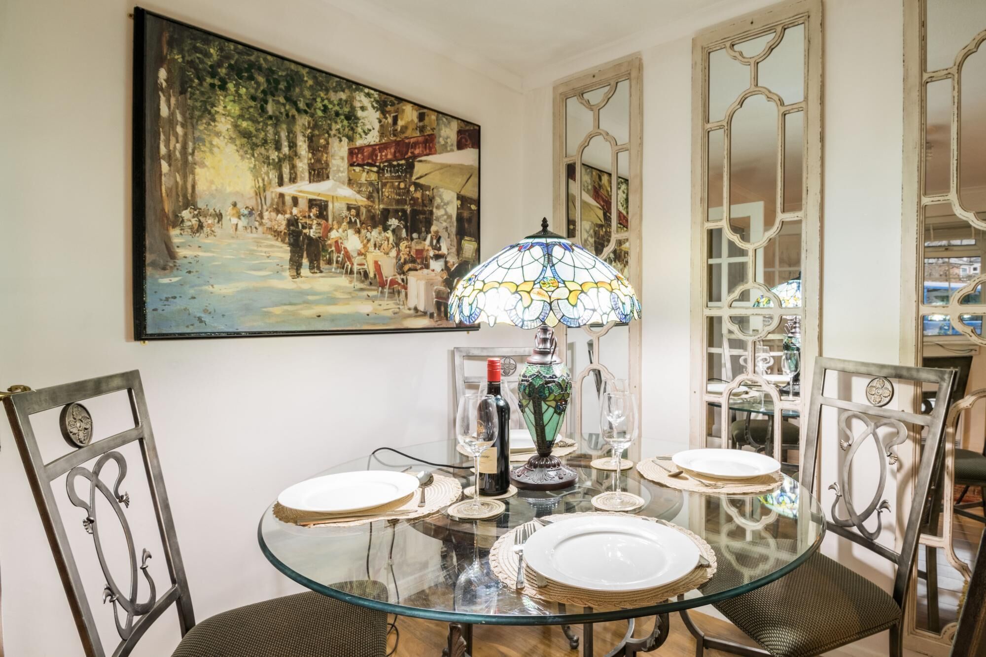 A dining area with a glass table, four chairs, a stained glass lamp, and a painting on the wall.