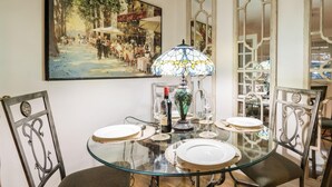 A dining area with a glass table, four chairs, a stained glass lamp, and a painting on the wall.