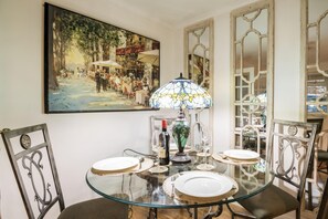 A dining area with a glass table, four chairs, a stained glass lamp, and a painting on the wall.