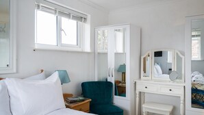 Ein Schlafzimmer mit einem Bett, einem Nachttisch, einem petrolfarbenen Sessel, einer weißen Kommode mit Spiegel und einem Fenster mit Jalousien.