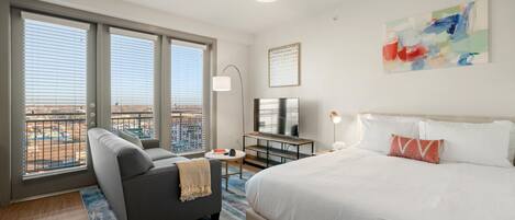 A hotel room with a large bed, a sofa, a TV, and a view of the city through large windows.