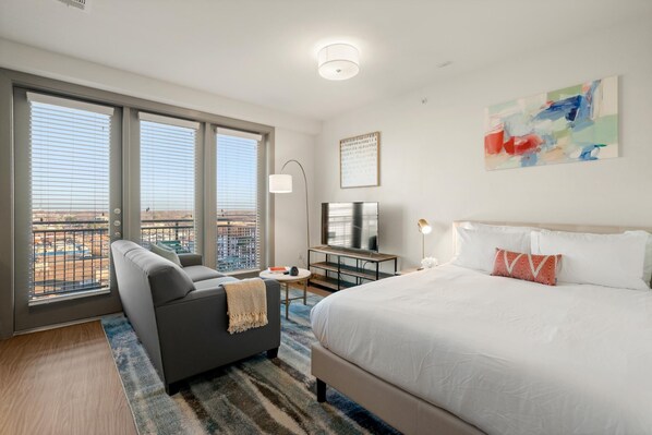 A hotel room with a large bed, a sofa, a TV, and a view of the city through large windows.
