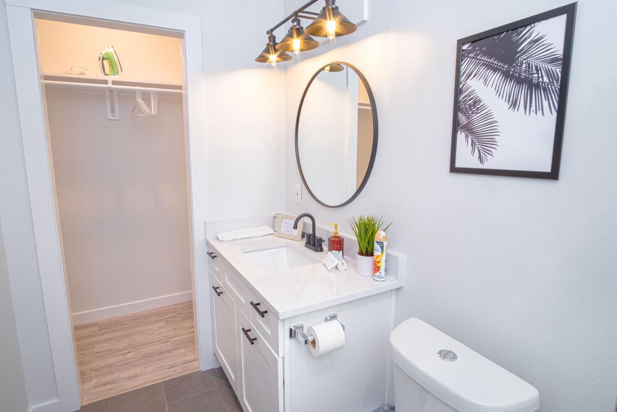 A modern bathroom with a white sink, mirror, and toilet. There are three pendant lights above the sink and a framed leaf artwork on the wall.