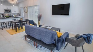 A modern living room with a sofa, a dining table, and chairs. There is a kitchen area with a microwave and cabinets. A flat-screen TV is mounted on the wall. - Brand NEW Stylish 3BR 2BA Near Exciting Downtown (San Antonio)