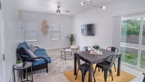 A modern living room with a sofa, coffee table, and dining table. There is a ceiling fan, a TV mounted on the wall, and a large window with blinds. - Brand NEW 4 Stylish 3BR Near Exciting Downtown (San Antonio)