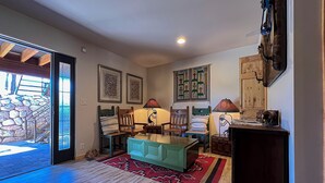 Een ruime woonkamer met een houten vloer, een groene bank, een eettafel en stoelen. Twee wandlampen en kunstwerken in lijsten zijn zichtbaar. Een glazen schuifdeur leidt naar een buitenruimte met een stenen muur en een houten trap.