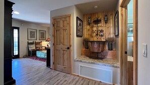 Een ruime woonkamer met een houten vloer, een grote houten deur en een keukenhoek met een koperen pan op het aanrecht.