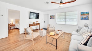 Un salon moderne avec un plancher en bois, un ventilateur de plafond, un téléviseur à écran plat et une grande fenêtre équipée de stores.
