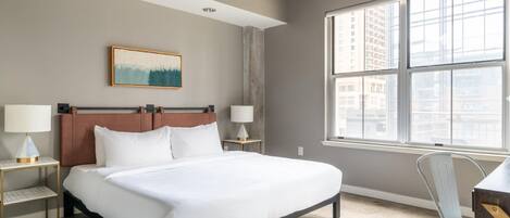A hotel room with a large bed, two bedside tables with lamps, a chair, a window with a city view, and a painting on the wall.