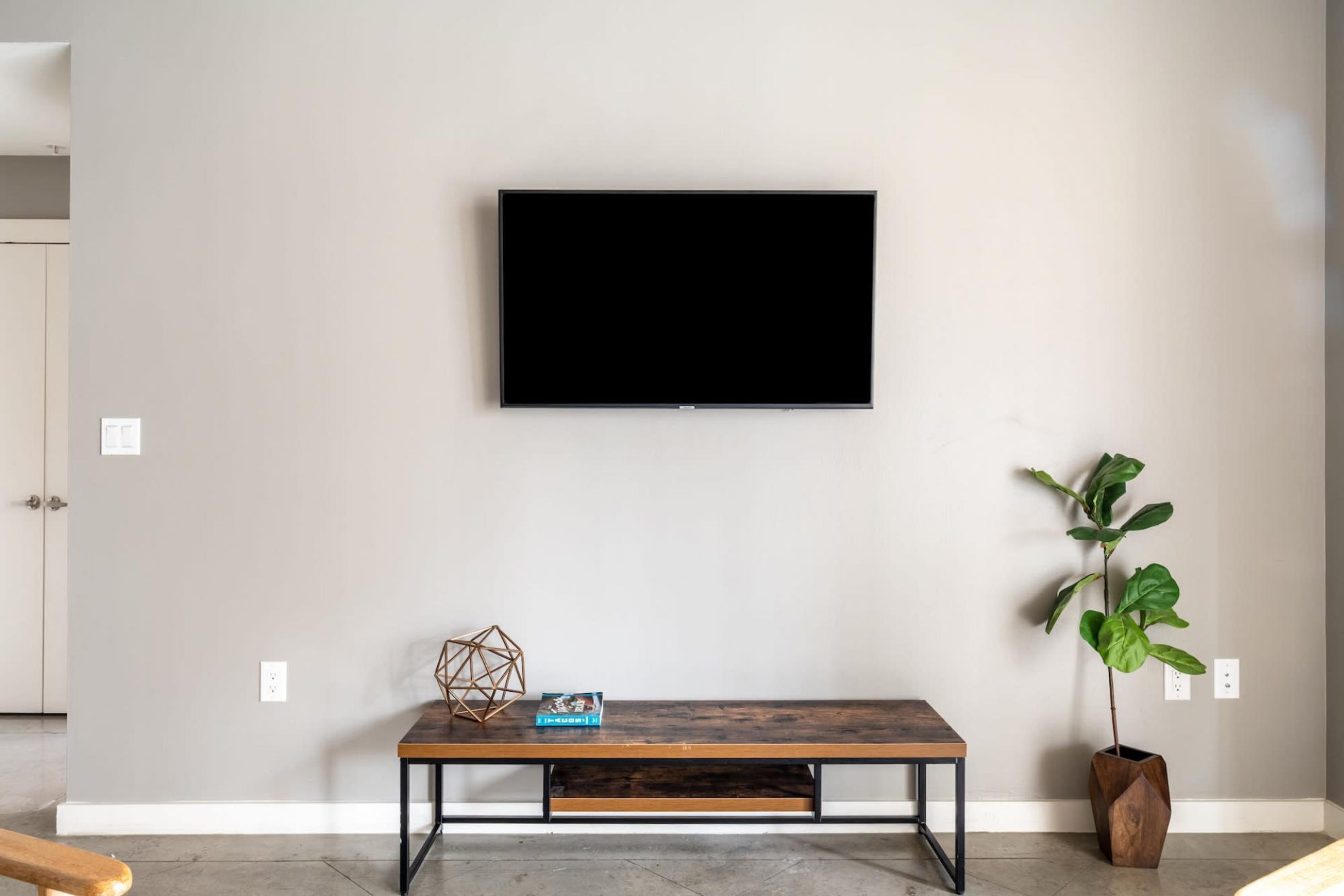 A modern living room with a flat-screen TV mounted on the wall, a wooden bench with a geometric decorative piece, and a potted plant.