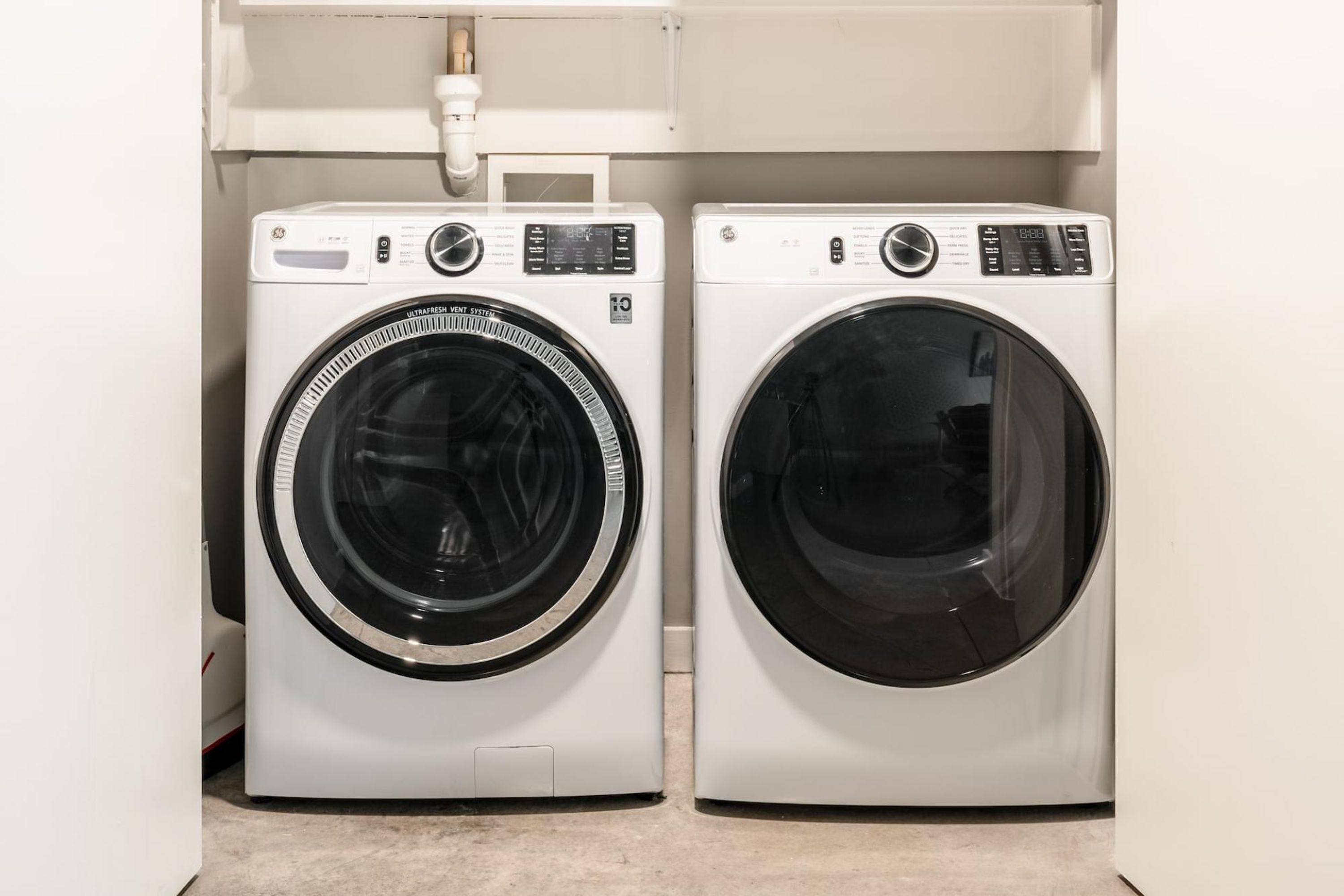 Two front-loading washing machines in a laundry room with a white wall and a visible pipe above them.