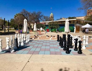 Un jeu d’échecs installé sur une terrasse carrelée, avec des parasols et un bâtiment en arrière-plan.